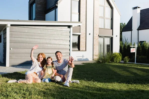 Щасливі батьки з махаючими руками і дочкою, сидячи на газоні і дивлячись на камеру біля будинку — стокове фото