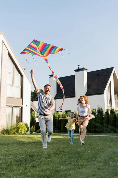 Comprimento total de feliz mãe e filha correndo perto do pai voando pipa no gramado perto de casas — Fotografia de Stock