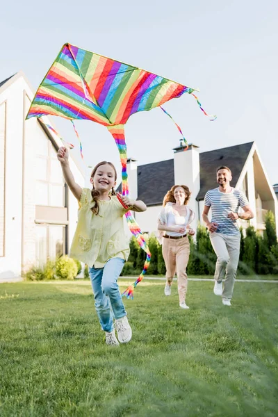 Glückliche Tochter blickt beim Drachenfliegen mit verschwommenen Eltern im Hintergrund in die Kamera — Stockfoto