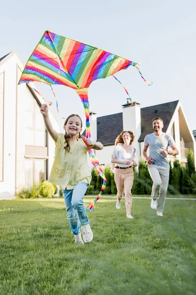 Feliz hija mirando a la cámara mientras vuela cometa con los padres borrosos en el fondo - foto de stock