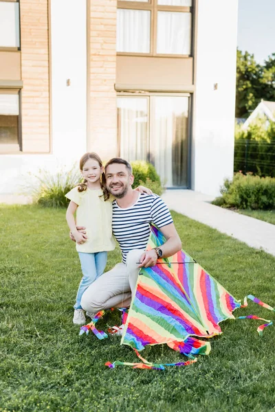 Padre feliz con la hija abrazando cometa y mirando a la cámara en el césped con la casa en el fondo - foto de stock