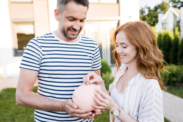 Sourire rousse femme mettre pièce de monnaie dans tirelire près de mari sur fond flou — Photo de stock