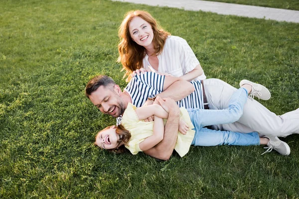 Glücklich rothaarige Frau blickt in die Kamera neben Mann kitzelt Mädchen, während sie auf dem Rasen liegt — Stockfoto