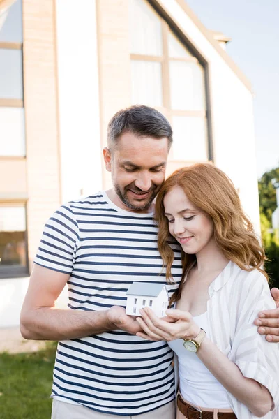 Lächelndes Paar umarmt sich und blickt auf Hausstatuette auf verschwommenem Hintergrund — Stockfoto