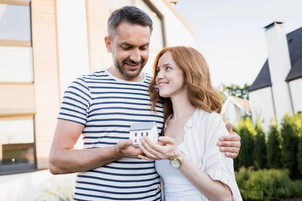 Smiling husband with statuette of house hugging wife on blurred background — Stock Photo