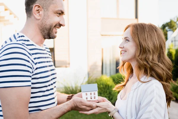 Vista lateral de la feliz pareja mirándose mientras sostiene la estatuilla de la casa sobre un fondo borroso - foto de stock