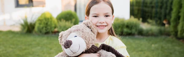 Porträt eines lächelnden Mädchens mit Teddybär, das auf verschwommenem Hintergrund in die Kamera blickt, Banner — Stockfoto