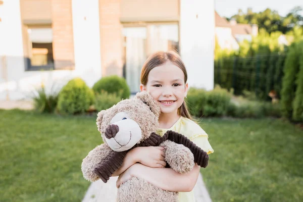 Menina feliz com ursinho olhando para a câmera com casa embaçada no fundo — Fotografia de Stock