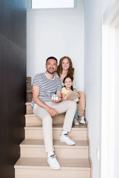 Familia feliz con oso de peluche y estatuilla de la casa mirando a la cámara mientras está sentado en las escaleras en casa - foto de stock