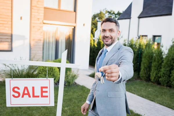 Happy broker looking at camera and standing near sign with sale lettering with blurred keys on foreground — Stock Photo