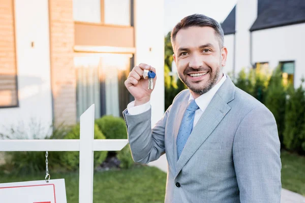 Happy broker showing keys while looking at camera on blurred background — Stock Photo
