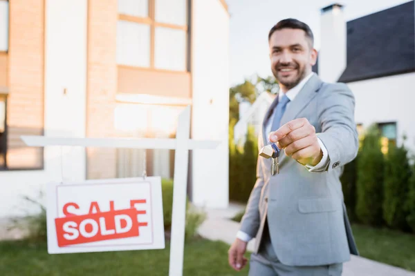 Smiling broker looking at camera while showing keys near sign with sold lettering on blurred background — Stock Photo