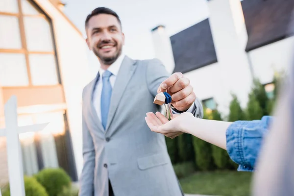 Sonriente agente de bienes raíces dando llaves a la mujer con casas borrosas en el fondo - foto de stock