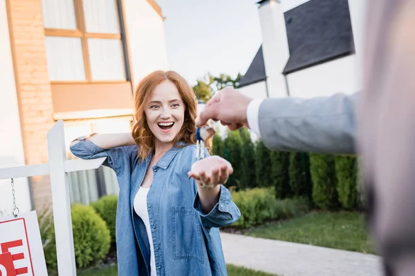 Femme rousse excitée avec bouche ouverte prenant les clés du courtier avec la main floue au premier plan — Photo de stock