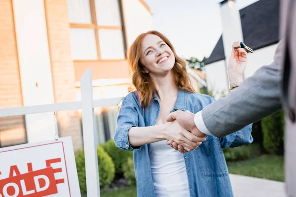 Glückliche rothaarige Frau mit Schlüsseln beim Händeschütteln mit Makler in der Nähe des Schildes mit verschwommenen Häusern im Hintergrund — Stockfoto