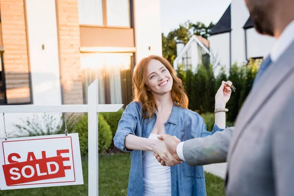 Joyeux rousse femme avec des clés serrant la main avec courtier près signe avec lettrage vendu au premier plan flou — Photo de stock