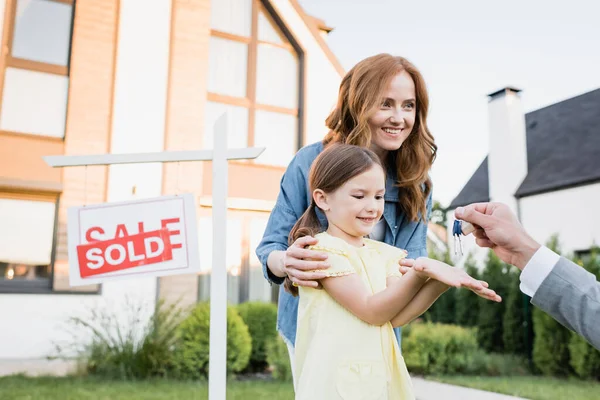 Happy mother hugging daughter taking keys from broker near sign with sold lettering on blurred background — Stock Photo