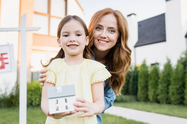 Rothaarige Mutter umarmt Tochter mit Hausstatuette, während sie in die Kamera schaut, mit verschwommenen Häusern im Hintergrund — Stockfoto