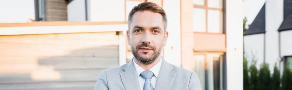 Confident bearded broker looking at camera with blurred houses on background, banner — Stock Photo