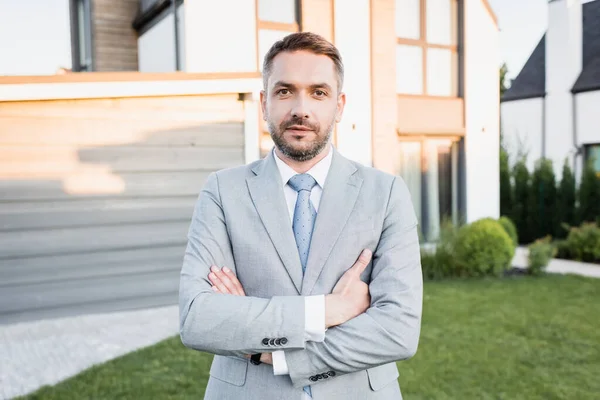 Selbstbewusster Makler mit verschränkten Armen blickt in die Kamera mit verschwommenen Häusern im Hintergrund — Stockfoto