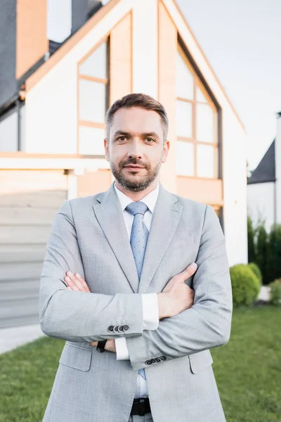 Front view of confident broker with crossed arms looking at camera with blurred house on background — Stock Photo