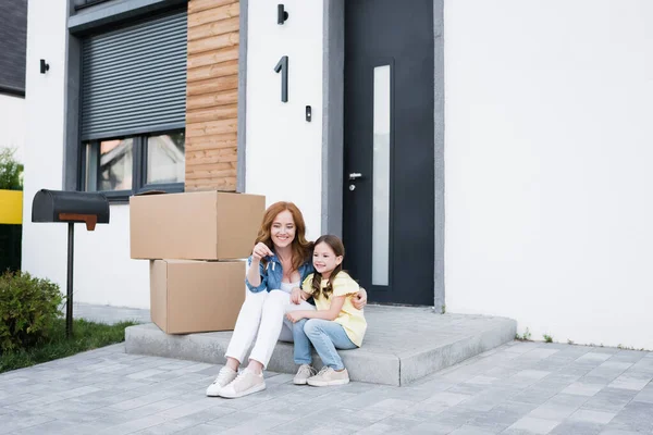 Mujer pelirroja feliz mirando a la llave y abrazando a su hija mientras está sentado en la puerta cerca de cajas de cartón - foto de stock