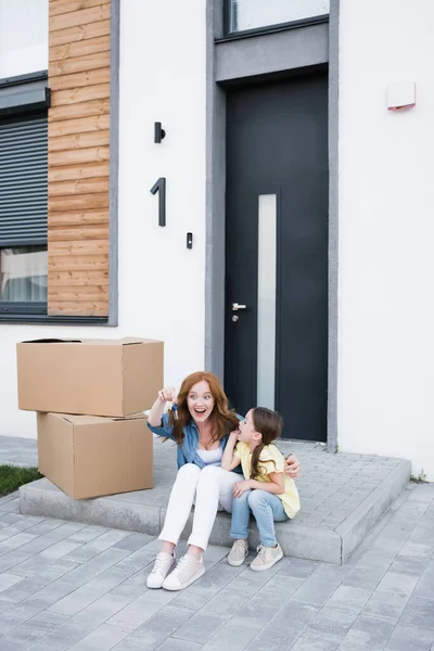 Aufgeregte Mutter und Tochter mit geöffnetem Mund schauen auf Schlüssel, während sie vor der Haustür in der Nähe von Kartons sitzen — Stockfoto