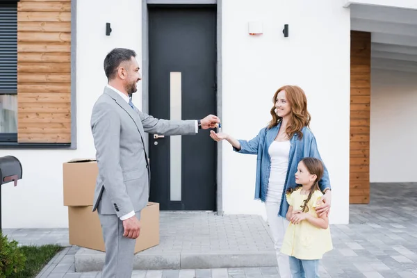 Happy broker giving key for woman with daughter near carton boxes on doorstep — Stock Photo