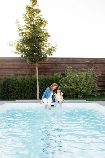 Fröhliche Mutter und Tochter sitzen mit Beinen im Schwimmbad im Hinterhof — Stockfoto