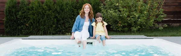 Allegro rossa madre guardando la fotocamera mentre seduto con figlia vicino alla piscina sul cortile, banner — Foto stock