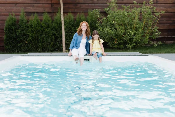 Lächelnde Mutter blickt in die Kamera, während sie neben ihrer Tochter in der Nähe des Swimmingpools im Hinterhof sitzt — Stockfoto