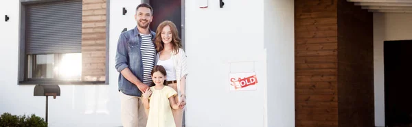 Famille heureuse regardant la caméra tout en se tenant ensemble près de la maison et signer avec lettrage vendu, bannière — Photo de stock