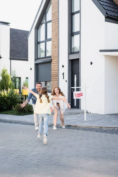 Back view of daughter with open arms running to happy parents near house and sign with sold lettering — Stock Photo