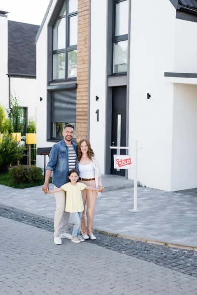 Familia feliz abrazando y mirando a la cámara mientras está de pie cerca de la casa y firmar con letras vendidas - foto de stock
