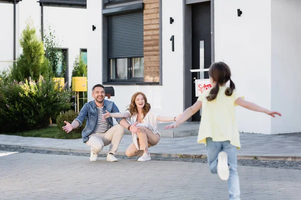 Happy parents squatting and looking at blurred daughter running on foreground — Stock Photo