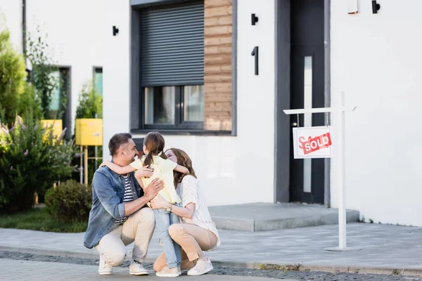 Felices padres en cuclillas y abrazando a la hija cerca de la casa en un fondo borroso - foto de stock