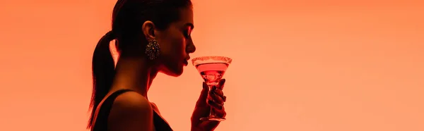 Side view of young woman holding margarita cocktail with ice cubes on orange, banner — Stock Photo