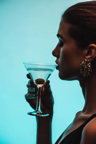 Side view of woman holding glass and drinking martini isolated on blue — Stock Photo