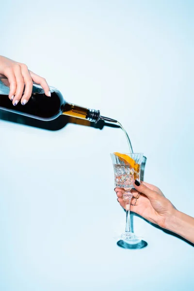 Abgeschnittene Ansicht einer Frau, die eine Flasche hält und Alkohol in ein Glas mit aufgeschnittener Orange auf Blau gießt — Stockfoto
