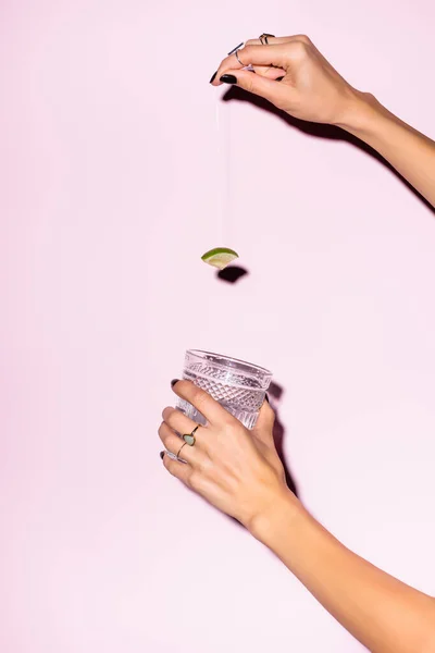 Обрезанный вид женщины, держащей лайм над стеклом с алкогольным напитком на розовом — стоковое фото