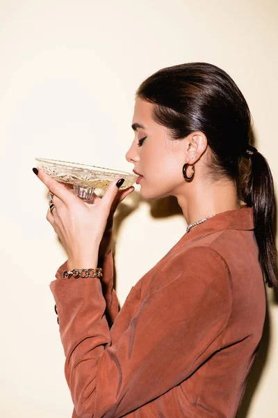 Vue latérale de la femme brune en blazer brun buvant du champagne à partir d'un verre sur blanc — Photo de stock