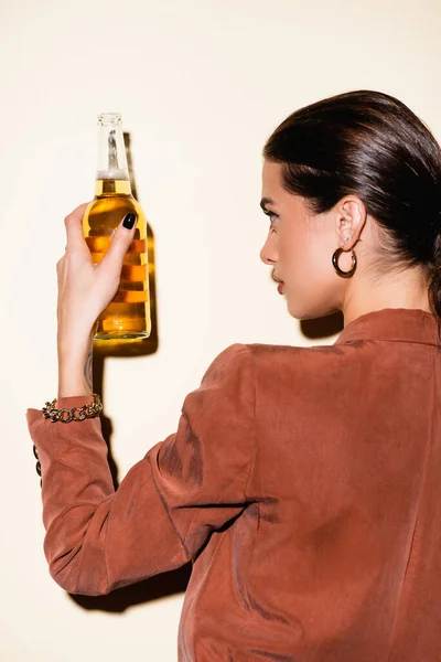 Morena mujer en chaqueta marrón mirando botella con cerveza en blanco - foto de stock