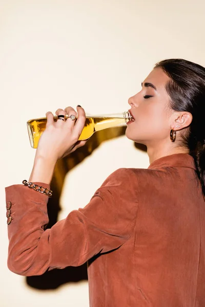 Vista lateral de mujer morena en chaqueta marrón bebiendo cerveza en blanco - foto de stock