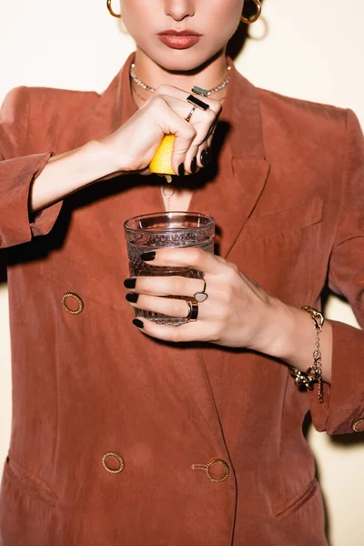 Vista recortada de mujer joven en chaqueta marrón exprimiendo limón en vidrio con cóctel de alcohol en blanco - foto de stock