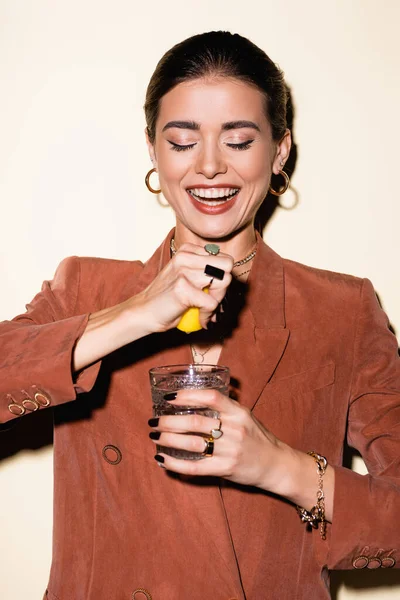 Mujer feliz en blazer marrón exprimiendo limón en vidrio con cóctel de alcohol en blanco - foto de stock