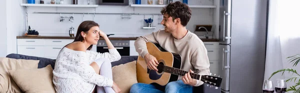 Sorrindo homem tocando guitarra acústica perto da namorada e copos de vinho em casa, banner — Fotografia de Stock