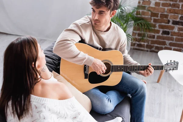 Joven tocando la guitarra acústica y mirando a su novia en primer plano borroso - foto de stock