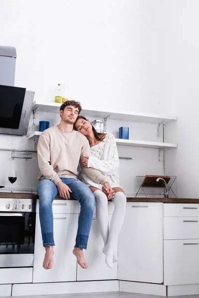 Junges Paar hält Händchen, während es mit geschlossenen Augen bei einem Glas Wein auf einer Arbeitsplatte in der Küche sitzt — Stockfoto