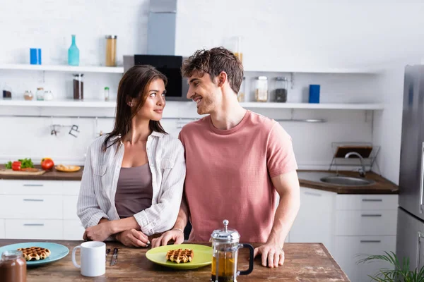Sorrindo homem de pé perto da namorada, waffles e chá na mesa da cozinha — Fotografia de Stock