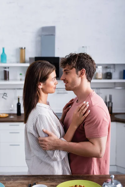 Vista laterale della giovane coppia che si abbraccia vicino al tavolo della cucina in primo piano sfocato — Foto stock